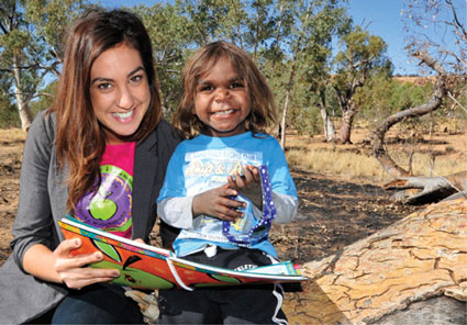 A smiling young woman with a smiling indigenous child looking up from a book they are reading